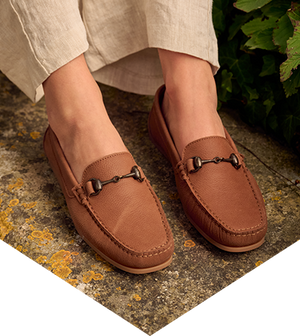 Brown loafers worn by a person on a stone surface with greenery in the background