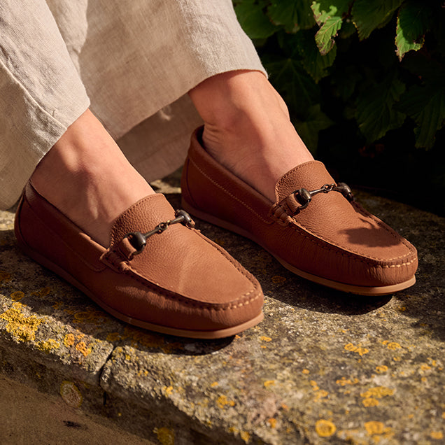 Dubarry Brown loafers with decorative straps worn by a person sitting on a stone surface with greenery in the background.