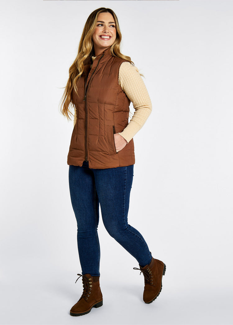 On model image of a pair of Dubarry Strokestown walnut brown leather women's short boots with brown padded collar, leather finger pull at the back and full laced up front with ski hooks. She is also wearing a brown quilted gilet and denim honeysuckle jeans