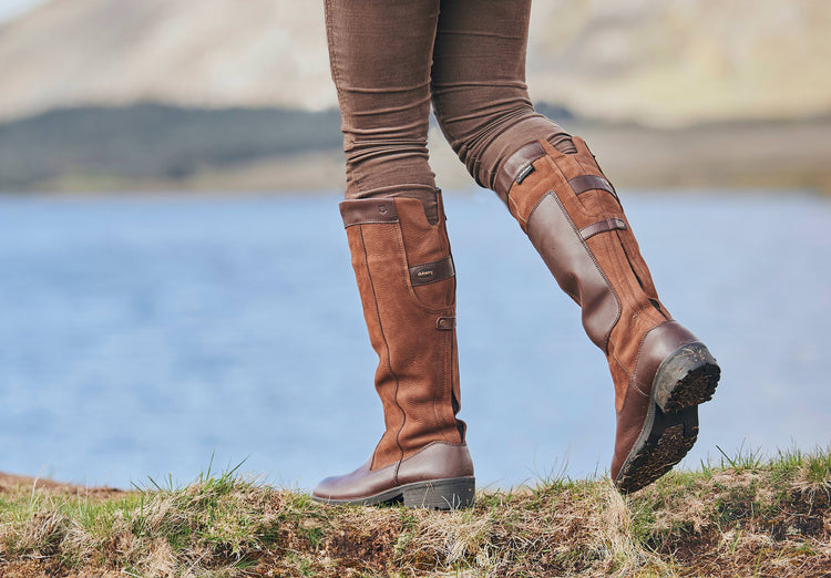 Image of a model wearing A pair of walnut brown Dubarry Clare leather country boots, knee high length with leather protection pad on inside leg beside a lake