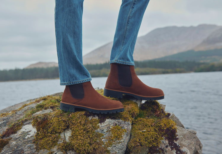 Outdoor set on model image of a woman wearing a pair of Dubarry Clonliffe Chelsea boots for women, walnut coloured leather chelsea boots with ridged elasticated side gussets and finger pull at the back. Model is standing on a boulder next to a body of water.