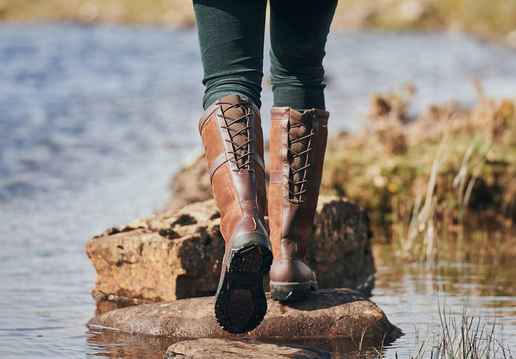 On model image of Pair of Dubarry ladies Glanmire adjustable calf country boot,  walnut brown knee high leather heeled boot with elasticated back and laced top. Model is walking along rocks jutting out of the water in a lake.