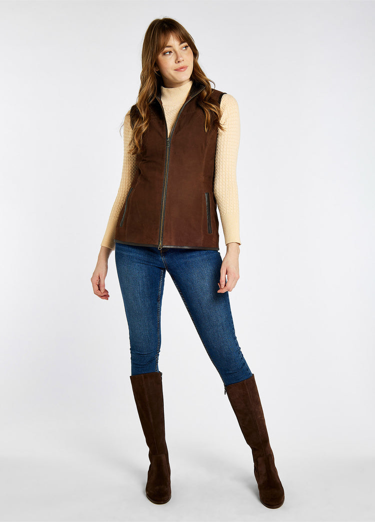 A model wearing Dubarry Kingston leather gilet zipped up, the walnut brown coloured gilet has pockets and a leather trim The model is also wearing a cream jumper, blue jeans and brown boots