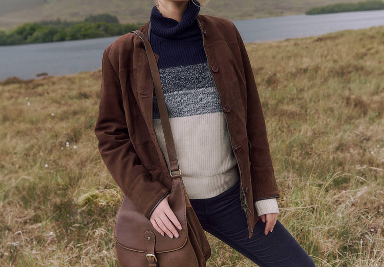 Oudoor image of a Model wearing a Dubarry Joyce women's mid length walnut brown coloured leather jacket with buttons. She is also wearing a knitted jumper that is navy, cream and grey.