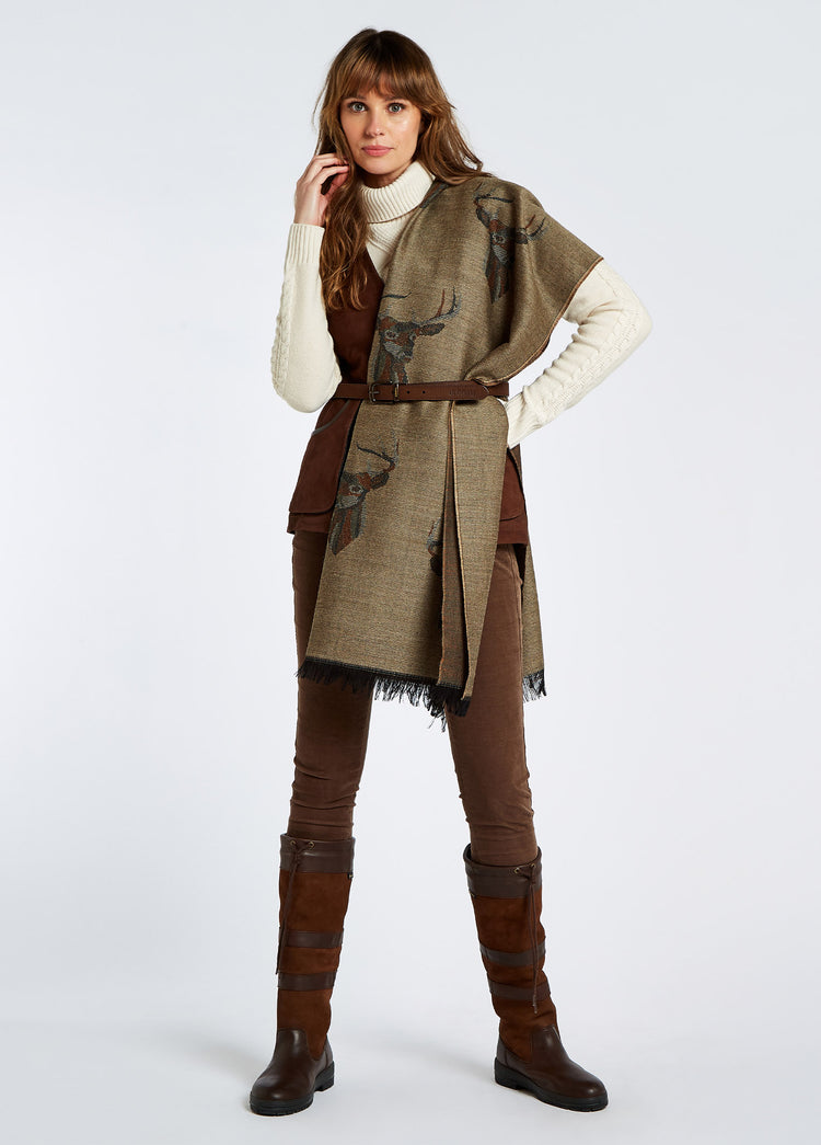 A woman modelling Dubarry women's Heatherbell wool jacquard with unique design in the colour camel, draped over her shoulders. She is also wearing brown honeysuckle jeans, cream knitted jumper, brown gilet and brown country boots.