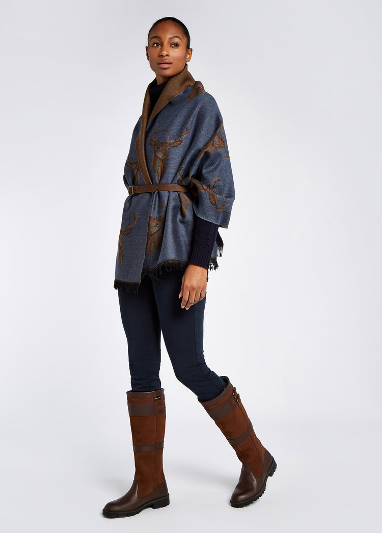 A woman modelling Dubarry women's Heatherbell wool jacquard with unique design in the colour indigo, draped around her torso. She is also wearing navy honeysuckle jeans, navy knitted jumper and brown country boots.