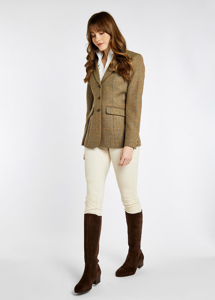 A woman modelling Dubarry women's Darkhedge Tweed Burren Jacket with stripe sleeve lining, buttons and single back vent. The model is also wearing white pants and brown boots.