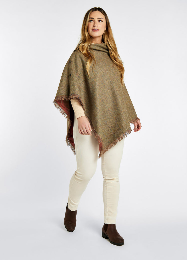A woman modelling Dubarry women's Hazelwood Tweed Burren Poncho with brass coloured buttons and 100% wool. She is also wearing white pants and brown boots . 