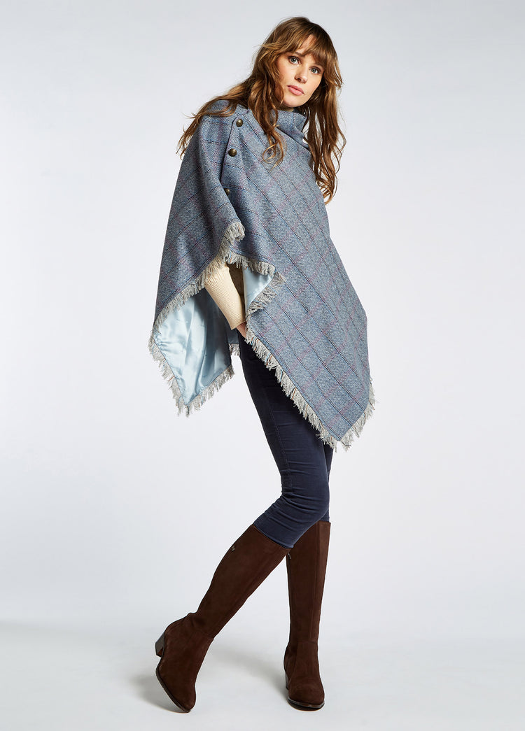 A woman modelling Dubarry women's Hazelwood Tweed Denim Haze Poncho. A fully lined poncho with functional button detail on one side. She is also wearing jeans and boots.