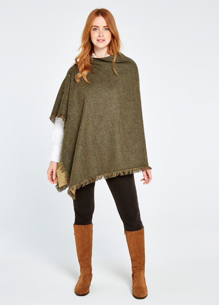 A woman modelling Dubarry women's Hazelwood tweed Heath Poncho with brass coloured buttons and 100% wool. She is also wearing black honeysuckle jeans and brown boots.