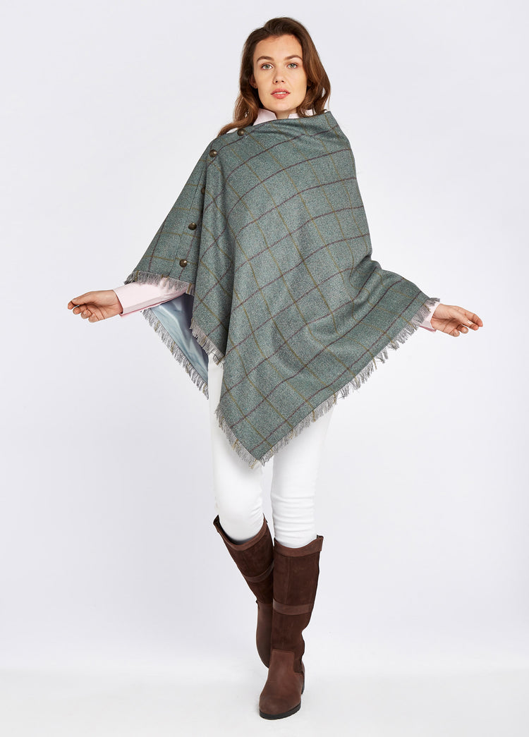 Woman modelling Dubarry Hazelwood Sorrel Poncho with smart brass coloured buttons and fabric fringe. She is also wearing white jeans and brown boots.