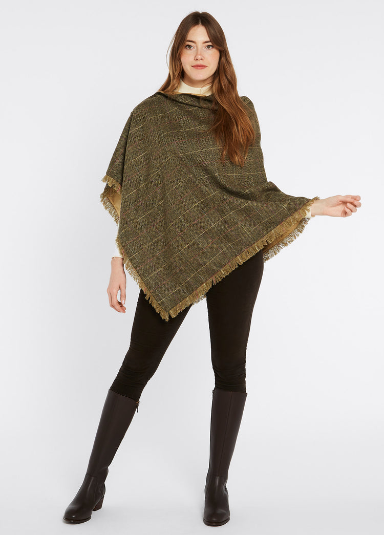 A woman modelling Dubarry women's Hazelwood tweed thistle Poncho with brass coloured buttons and 100% wool. She is also wearing brown pants and brown boots.