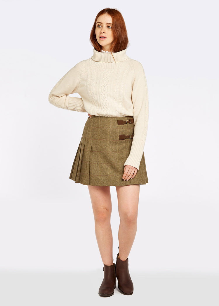 A woman modelling Dubarry women's Blossom Tweed Elm Skirt with two buckled leather tabs and stitched-down knife pleats. The model is also wearing a jumper and boots.