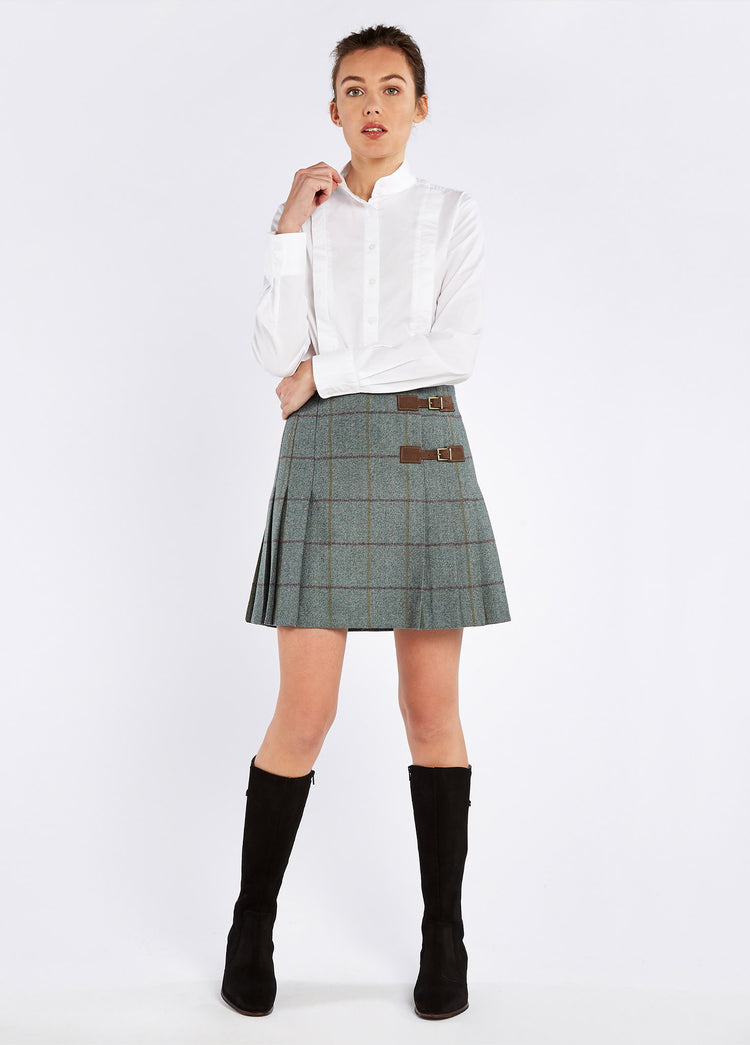 A woman modelling Dubarry women's Blossom Tweed Sorrel Skirt with two buckled leather tabs and stitched-down knife pleats. The model is also wearing a white shirt and black boots.