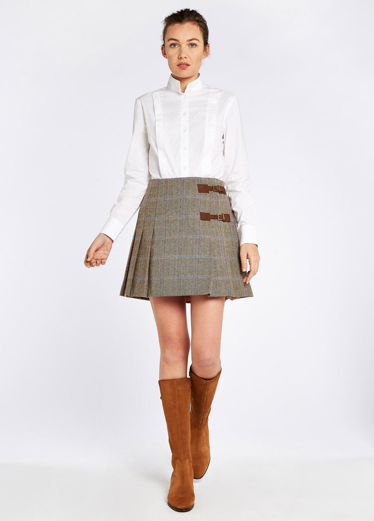 Woman modelling Blossom Tweed Skirt in the colour WoodRose. A pleated skirt with a polo and light knit and two buckled leather tabs. She is also wearing a white shirt and boots.