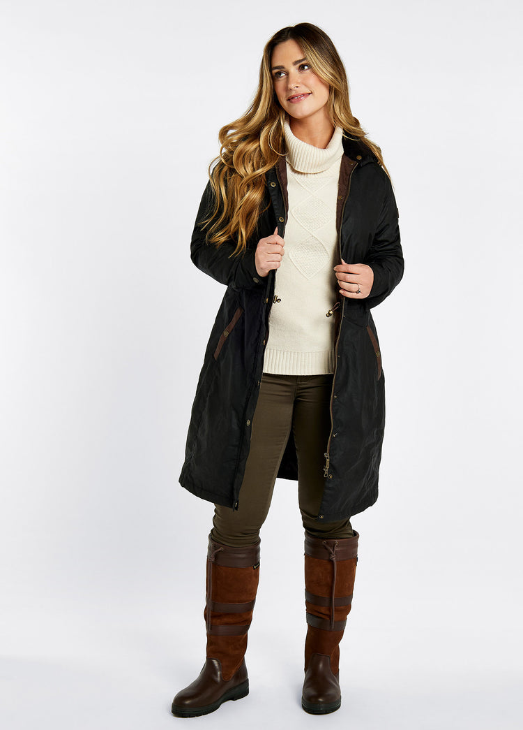 A female model wearing a black Dubarry Redington Wax coat, longer length coat with  full zip. Model is wearing a light coloured jumper underneath, brown jeans and boots