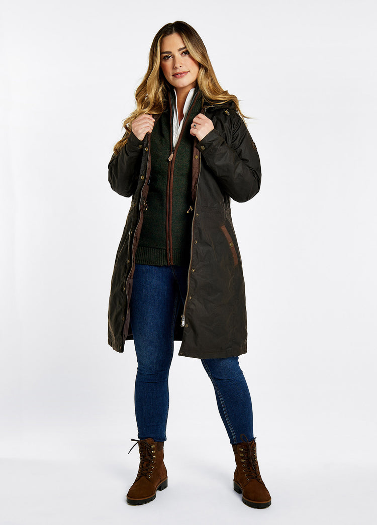 A female model wearing a black brown Dubarry Redington Wax coat, longer length coat with  full zip. Model is wearing a light coloured jumper underneath, light jeans and boots