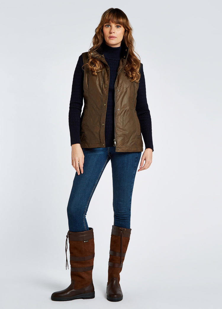A female model wearing a Dubarry Pakenham wax gilet jacket, ginger brown colour with buttons and zip. The model is also wearing a dark jumper underneath, blue jeans and boots
