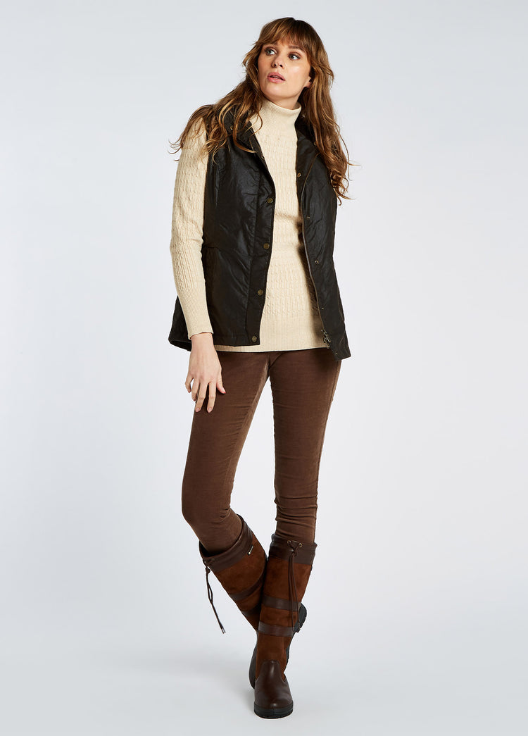 A female model wearing a Dubarry Pakenham wax gilet jacket, black colour with buttons and zip. The model is also wearing a cream jumper underneath, brown jeans and boots