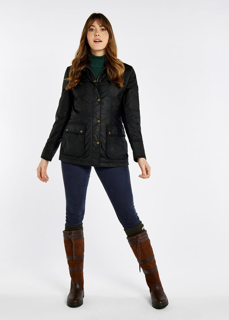 A female model wearing a black Dubarry Munsboro wax jacket, high collared jacket with golden buttons, pockets and full zip. The model is also wearing  a green top underneath, blue jeans and brown boots