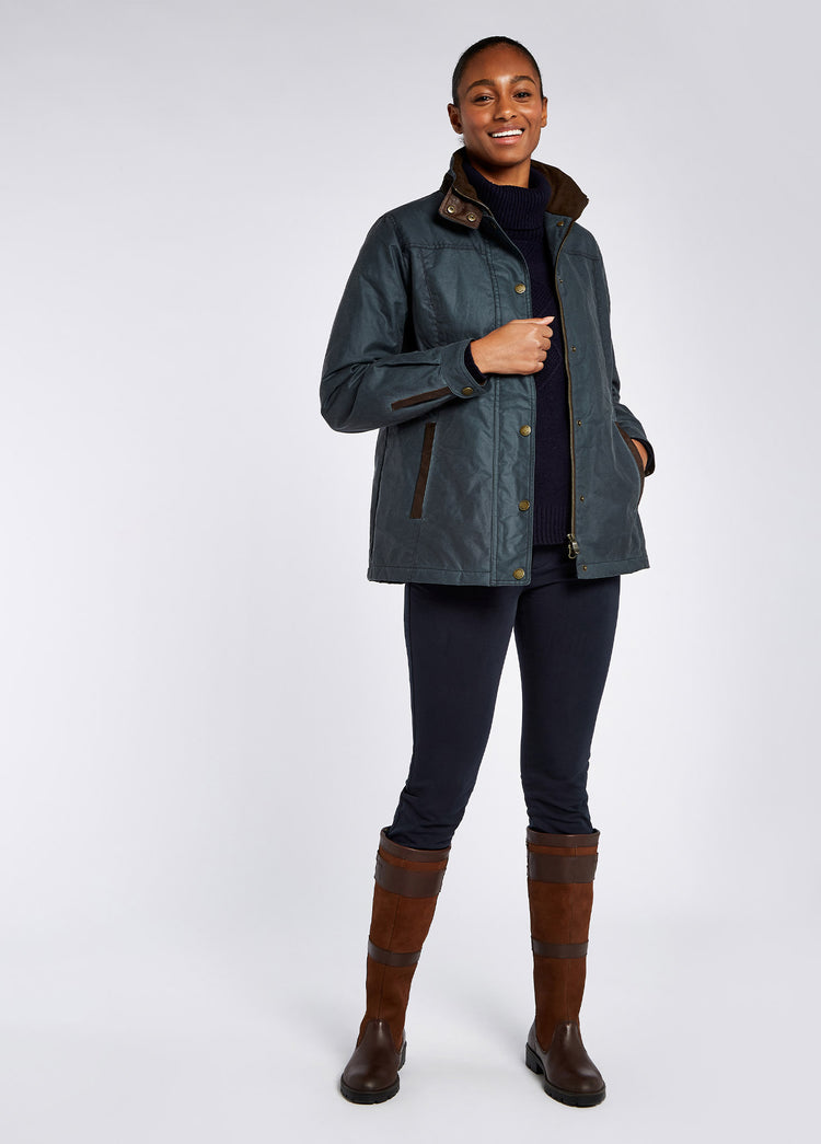 A female model wearing an opened Dubarry Mountrath Waxed jacket, dark pebble green blue coloured jacket with full zip and buttons, brown edged pockets, inner collar and brown inside collar. The model is also wearing a dark top underneath, dark jeans and brown boots