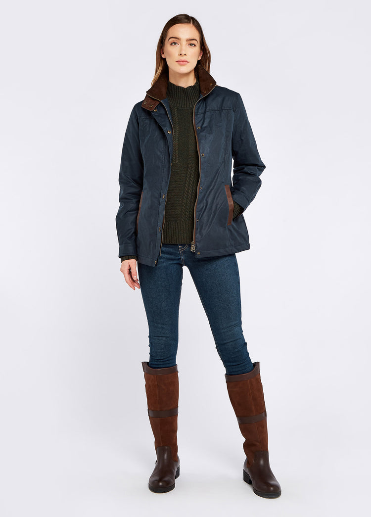 A female model wearing an opened Dubarry Mountrath Waxed jacket, ocean blue coloured jacket with full zip and buttons, brown edged pockets, inner collar and brown inside collar. The model is also wearing a dark top underneath, dark jeans and brown boots