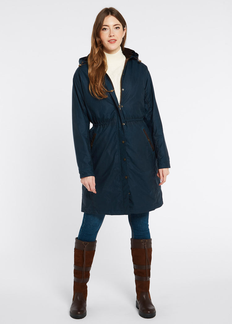 A female model wearing a storm blue Dubarry Redington Wax coat, longer length coat with  full zip. Model is wearing a cream coloured jumper underneath, light jeans and brown boots