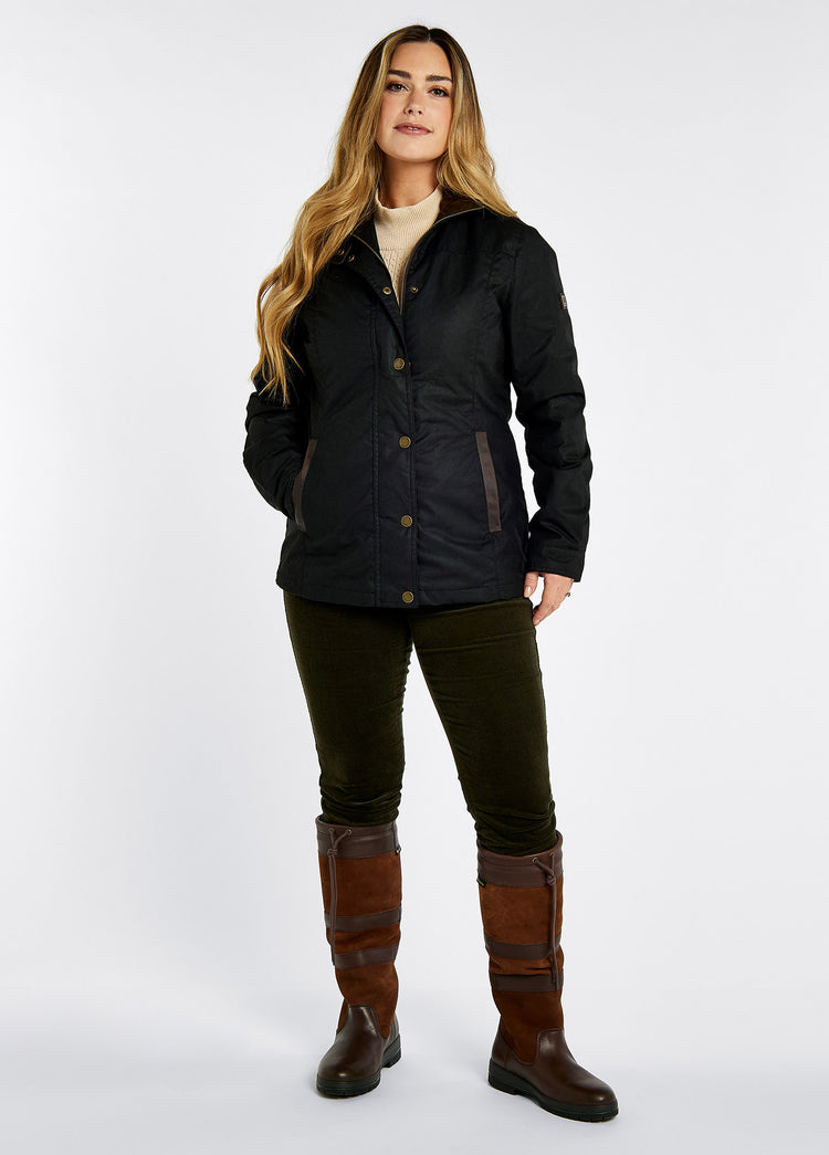 A female model wearing a Dubarry Mountrath Waxed jacket, black jacket with full zip and buttons, brown edged pockets, inner collar and brown inside collar. The model is also wearing a light top underneath, dark jeans and brown boots