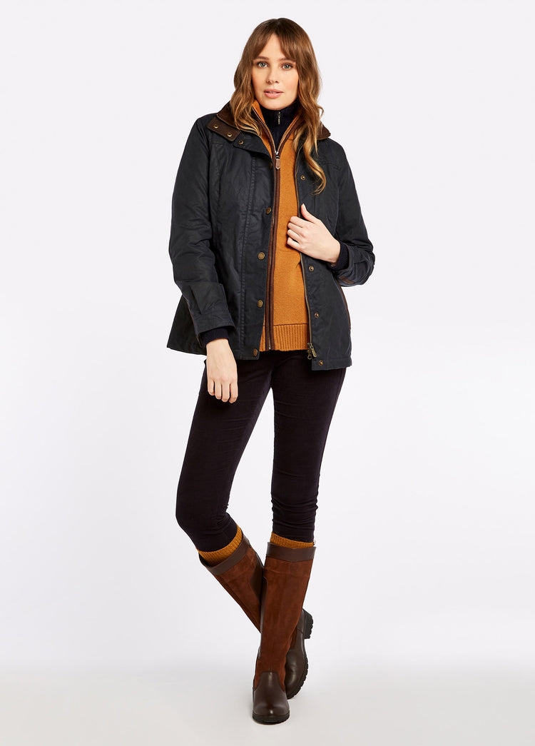 A female model wearing a Dubarry Mountrath Waxed Jacket, a warm winter jacket with vertical hip pockets, metal front zip and high collar The model has the jacket zipped fully down and is wearing an orange jumper, black pants and brown boots. 
