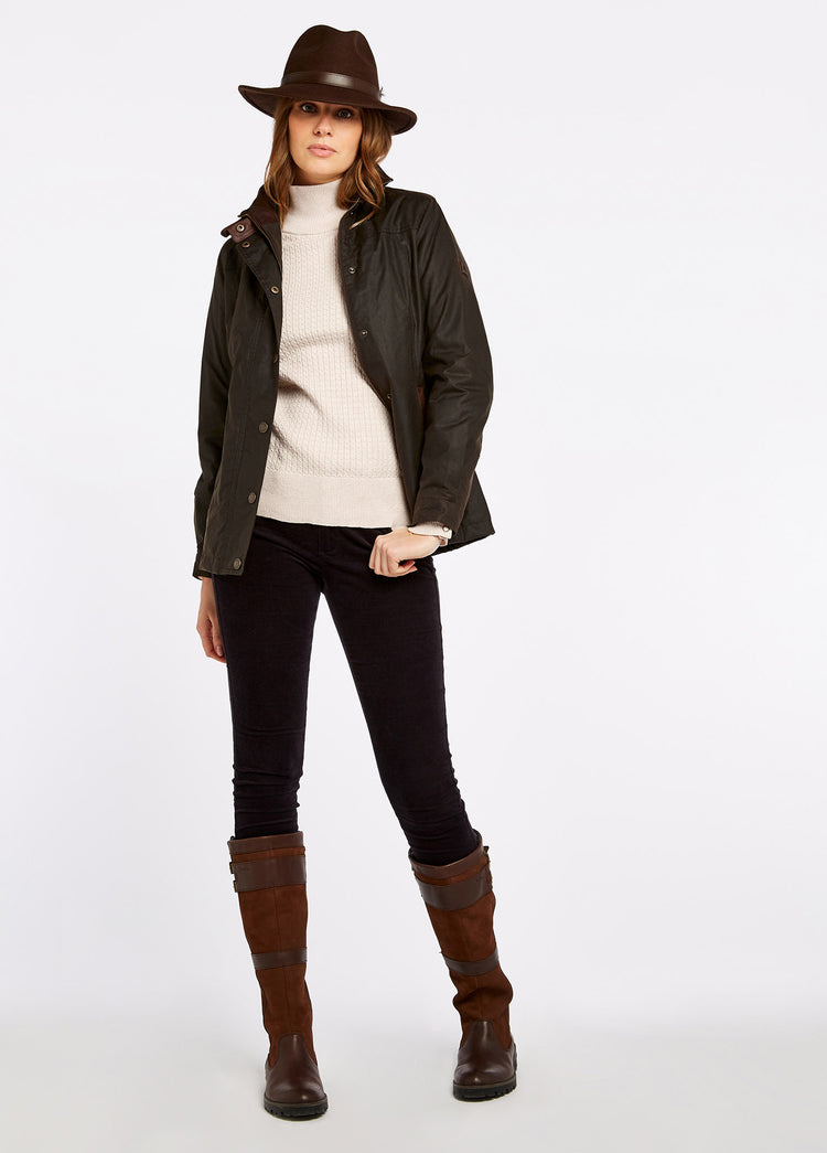 A female model wearing a Dubarry Mountrath olive brown coloured waxed jacket opened, with fill zip and buttons with vertical leather detail on the jacket cuffs. The model is also wearing a cream jumper underneat the jacket, a brown hat, blue jeans and brown boots