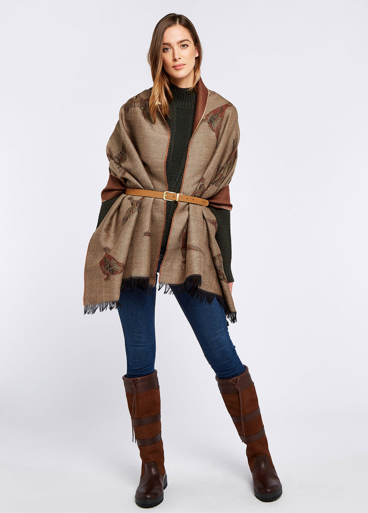 A woman modelling Dubarry women's Birchdale Tweed Taupe Stole draped over her shoulder and tied with a belt. She is also wearing a hat, blue jeans and brown boots.