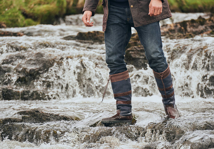 On Model image of a man wearing a pair of Dubarry Galway knee high leather country boots, in the colour navy/brown; standing in the banks of a river.