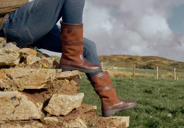 On model image of a pair of Dubarry Mens Kildare calf height leather Country Boot, walnut brown colour with model sitting on a boulder in a grassland.
