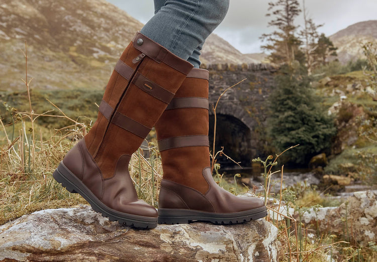 On Model image of a man wearing Dubarry knee-high walnut brown leather Wexford wider fitting Country boots with full zip standing on a boulder in a grassland area.