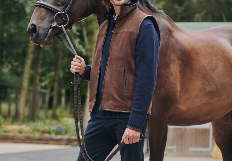Outdoor image of male model with a horse. Image is taken from the waist up view, modelling a Dubarry Dunhill walnut brown coloured gilet leather jacket with centre zip, the jacket is open and the man is wearing a navy jumper with a white shirt underneath and jeans.