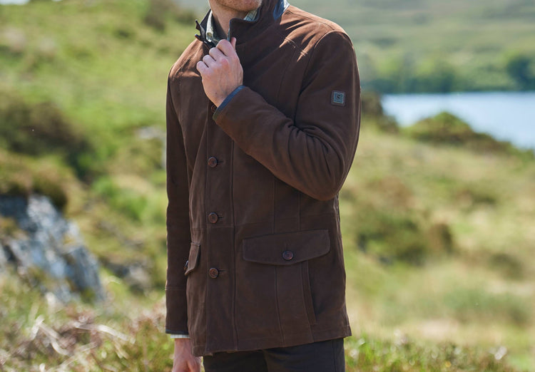 An outdoor image in a grassland of a male modelling Dubarry Moore Walnut coloured leather jacket , the jacket is closed, showcasing the stand up collar , zip, quilited lining, pockets and buttons.