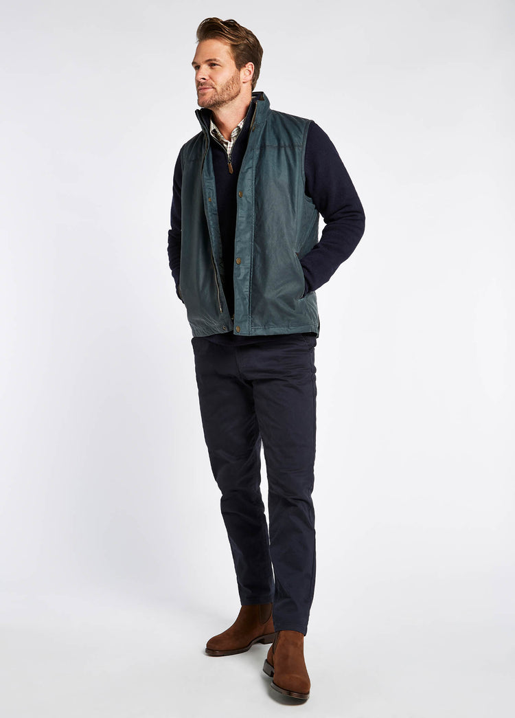 Male model wearing a Dubarry Mayfly wax gilet jacket for men. Dark pebble green blue coloured gilet with full zip opened, inside collar of the jacket is a lighter shade of brown with buttons along the zip. The model is also wearing a dark jumper and light shirt underneath the gilet, dark jeans and brown boots.
