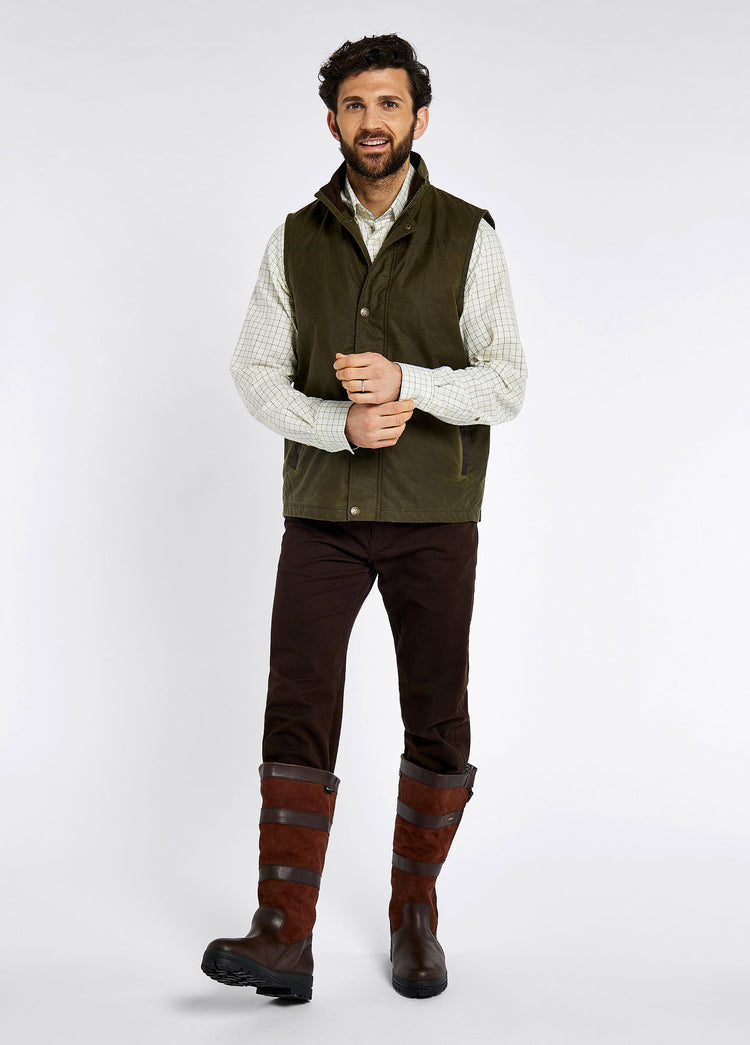 Male model wearing a Dubarry Mayfly wax gilet jacket for men, fennel gilet with full zip opened, inside collar of the jacket is a lighter shade of brown with buttons along the zip. The model is also wearing a light shirt underneath the gilet, dark jeans and brown boots.