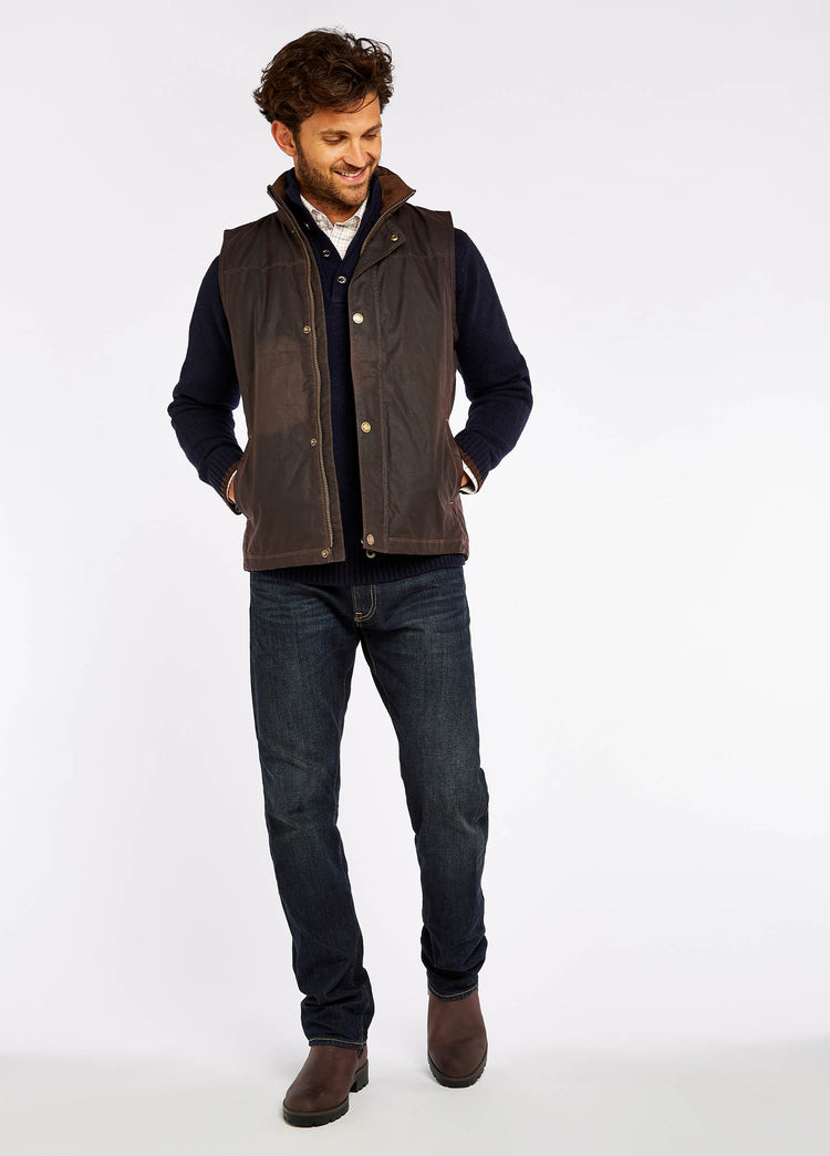 Waist up view of a male model wearing a Dubarry Mayfly wax gilet jacket for men. Java brown coloured gilet with full zip opened, inside collar of the jacket is a lighter shade of brown with buttons along the zip. The model is also wearing a dark jumper and light shirt underneath the gilet.