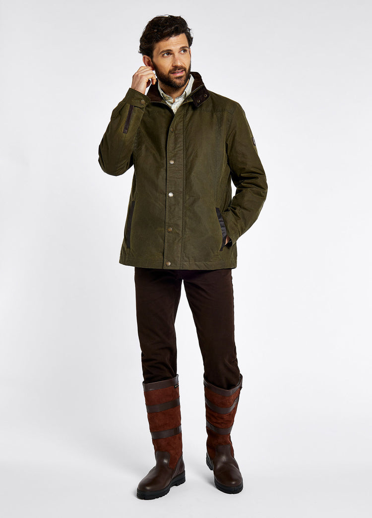 A male model wearing a Dubarry Carrickfergus waxed jacket open, fennel coloured with a standing collar and buttons along the full zip and lighter brown inside collar. The model is also wearing a dark jumper, light shirt, dark jeans and brown boots