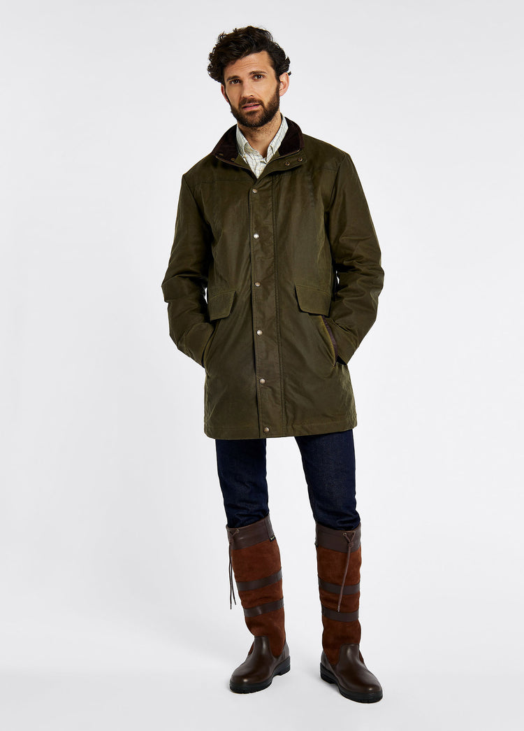 Male model wearing a men's Dubarry longer length wax jacket opened, fennel colour with full zip, buttons and pockets. The model is also wearing a dark jumper and white shirt undeneath with dark jeans and brown boots