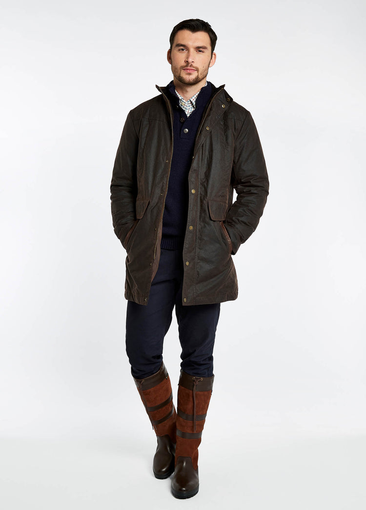 Male model wearing a men's Dubarry longer length wax jacket opened, java colour with full zip, buttons and pockets. The model is also wearing a dark jumper and white shirt undeneath with dark jeans and brown boots