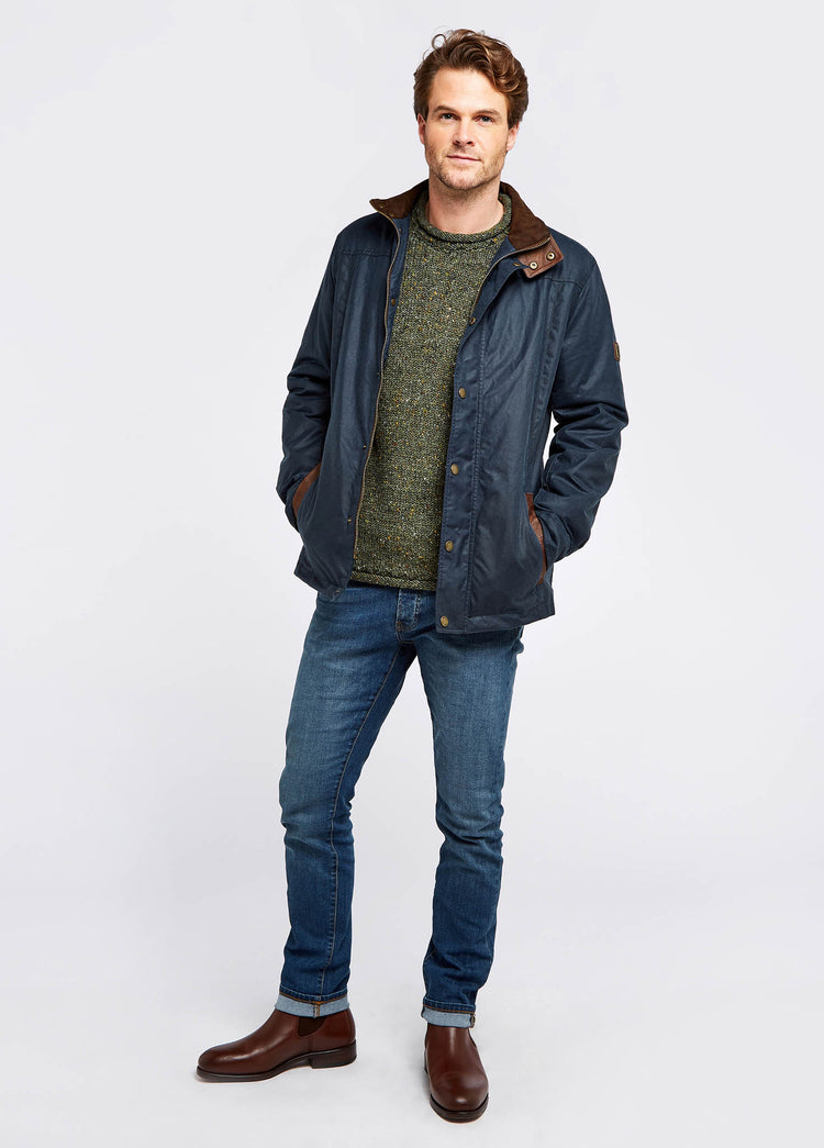 Waist up view of a male model wearing a Dubarry Carrickfergus waxed jacket, ocean blue coloured with a standing collar and buttons along the full zip, the model is wearing the jacket open showing the lighter brown inside collar and he is wearing a green jumper and blue jeans