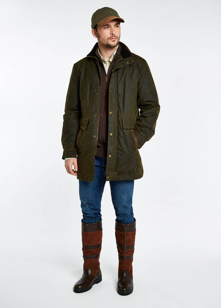 Male model wearing a men's Dubarry longer length wax jacket opened, olive colour with full zip, buttons and pockets. The model is also wearing a dark jumper and shirt undeneath with blue jeans and brown boots