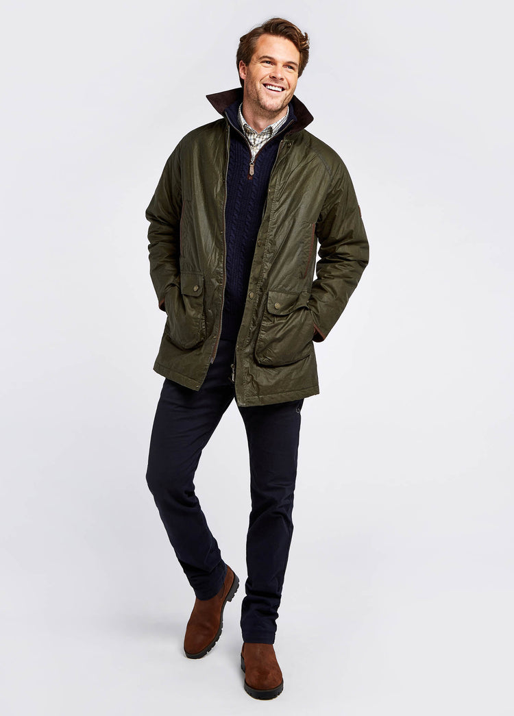A male model wearing a Dubarry Brunswick Wax Jacket, pine green coloured longer length jacket with a brown corduroy collar and full zip and buttons, there is multiple pockets on the jacket. The model also wears a jumper and shirt underneath the jacket, dark jeans and brown boots.