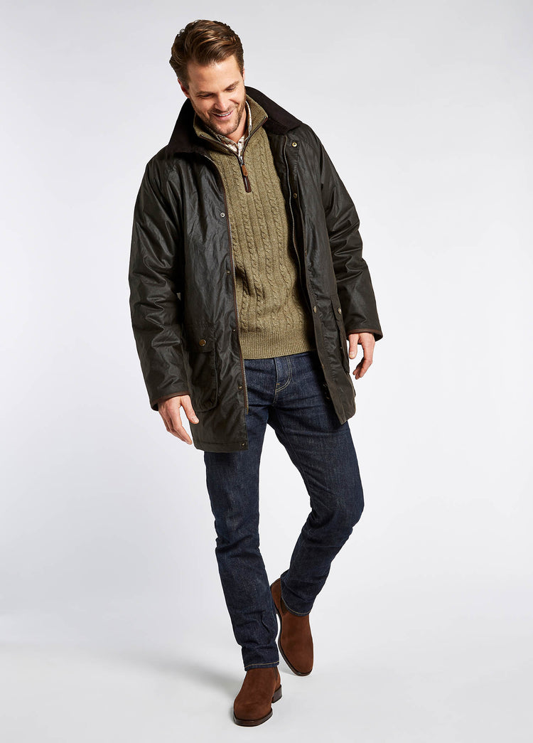 A male model wearing a Dubarry Brunswick Wax Jacket, midnight navy coloured longer length jacket with a brown corduroy collar and full zip and buttons. The model has his hands in the pockets of the jacket, there is multiple pockets on the jacket. The model also wears a jumper and shirt underneath the jacket, dark jeans and brown boots.