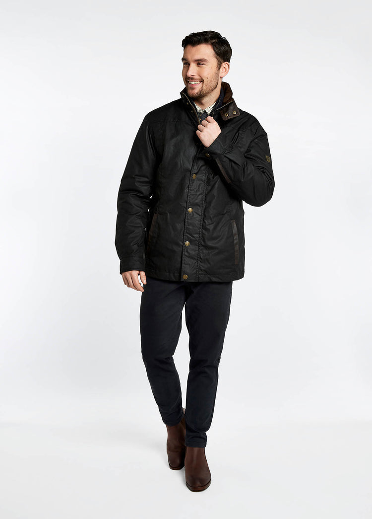 A male model wearing a black Dubarry Carrickfergus waxed jacket with a standing collar and buttons along the full zip and lighter brown inside collar. The model is also wearing a dark jumper, light shirt, dark jeans and brown boots