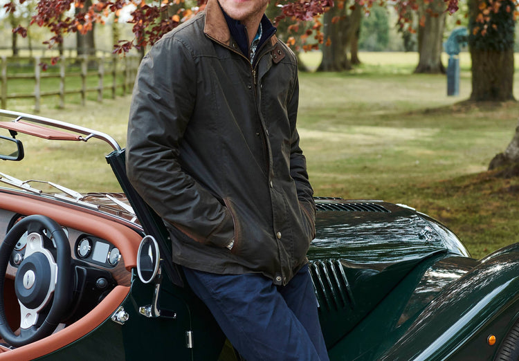 On model image of man sittin gon a vintage car. He is wearing A Dubarry Carrickfergus waxed jacket, olive coloured with a standing collar and buttons along the full zip and two  pockets. The model has his hands in the pockets and is also wearing dark jeans.